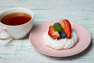 Pavlova cake with strawberries and blueberries and mint on a pink saucer and a cup of hot tea. Home confectionery. Old retro white and blue table.