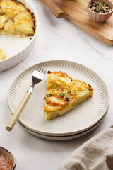 Slice of potato casserole with red onions, fresh thyme and rosemary on green checkered kitchen towel , wooden board with flavoring ingredients: pepper, salt, olive oil