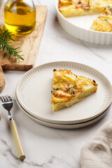 Slice of potato casserole with red onions, fresh thyme and rosemary on green checkered kitchen towel , wooden board with flavoring ingredients: pepper, salt, olive oil