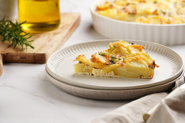 Slice of potato casserole with red onions, fresh thyme and rosemary on green checkered kitchen towel , wooden board with flavoring ingredients: pepper, salt, olive oil