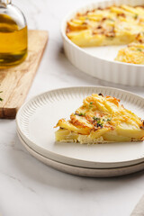 Slice of potato casserole with red onions, fresh thyme and rosemary on green checkered kitchen towel , wooden board with flavoring ingredients: pepper, salt, olive oil