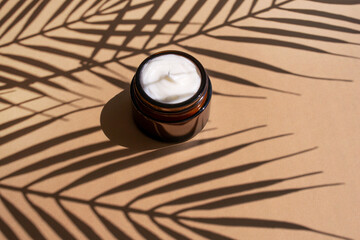 Cosmetic creme in dark glass jar on beige background. Organic moisturizing balm with palm leaves shadow.