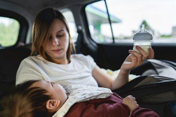 Adult caucasian woman female mother sitting on the back seat of her car holding formula or breast milk in baby bottle ready to feed her child while traveling on the road in summer day on holiday