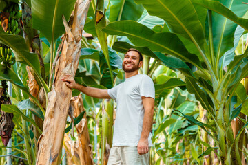 Happy man jungle portrait. Farmer agronomist or happy male tourist portrait near young palm banana trees on banana plantation, banana farm, tropical garden or rural farm in harvesting time.