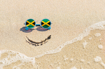 Fototapeta na wymiar A painted smile on the sand and sunglasses with the flag of Jamaica. The concept of a positive and successful holiday in the resort of Jamaica.