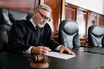 senior judge in robe and eyeglasses holding pen near paper and blurred gavel