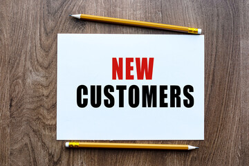 The text NEW CUSTOMERS is written in a notebook which lies on a wooden table. Lettering on a business or financial theme. Business concept.