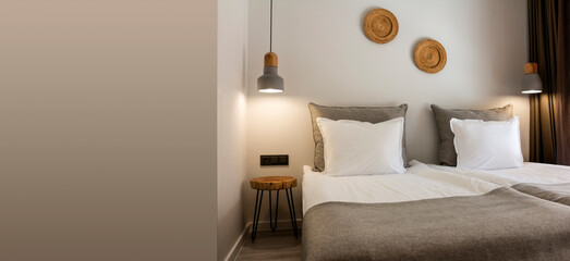 Bright cozy seating area with two single beds with white and gray linens. Stylish laconic lighting above the bedside table is attached. The wall is decorated with straw wicker plates. Banner.
