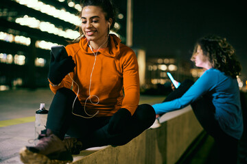 A smiling fit sportswoman sitting outside at night, relaxing and using phone.