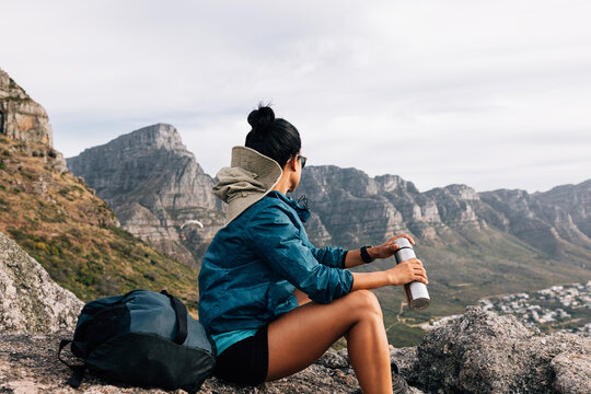 Female mountain climber looking at the view while sitting on a rock, holding a thermos