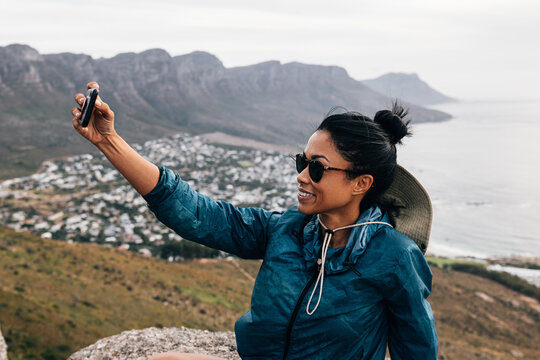 Young woman in sunglasses holding a smartphone and taking a selfie while taking a break during a mountain hike