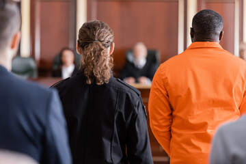 back view of accused african american man near guard and blurred jurors in courtroom