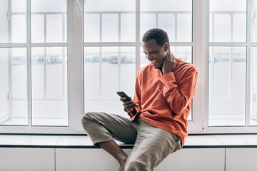 Positive African-American guy in stylish orange shirt looks out of window smiling with modern mobile phone in light room at home