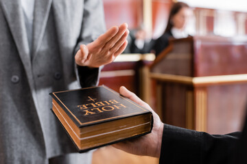 cropped view of witness giving swear on bible near blurred prosecutor in courtroom - obrazy, fototapety, plakaty