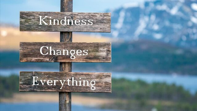 kindness changes everything text on wooden signpost outdoors in landscape scenery during blue hour. Sunset light, lake and snow capped mountains in the back.