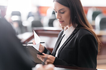 brunette prosecutor reading lawsuit near blurred judge in courtroom