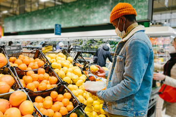 African-American man customer in warm jacket with disposable mask takes fresh lemon fruits from crate in supermarket side view