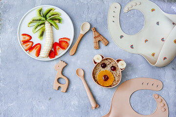 Funny bowl with oat porridge with bear faces made of fruits and berries on blue background. Food for kids idea, top view, copy space.