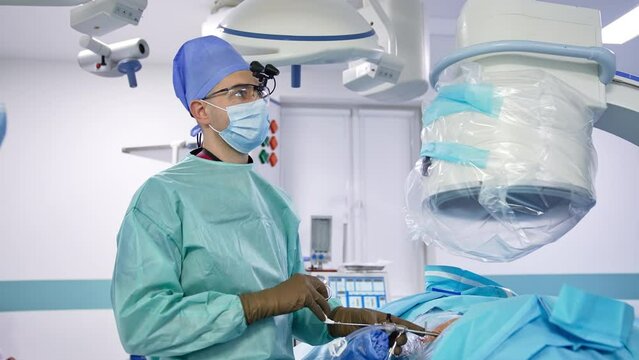 Middle-aged neurosurgeon looks straight at camera and speaks. Specialist pulls the instrument and gives it to assistant for cleaning.