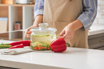 Woman with cooking pot of tasty dietary soup on table in kitchen