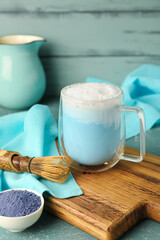 Wooden board with cup of blue matcha latte, powder and chasen on color wooden background
