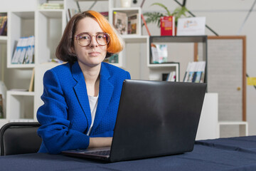 Young beautiful and stylish girl with multi-colored hair in glasses and blue business suit sits at laptop. Looks at camera.