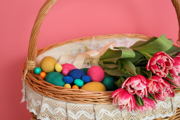 Wicker basket with Easter eggs and flowers on pink background, closeup
