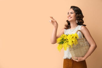 Beautiful young woman with mimosa flowers in bag pointing at something on color background