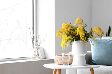 Vase with mimosa flowers on table and sofa in interior of living room