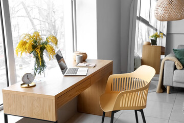 Vase with mimosa flowers and laptop on table in interior of room