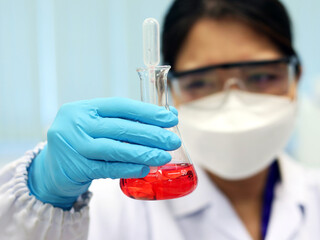 The scientist wears PPE and holds an Erlenmeyer flask laboratory, with red solvent solution from...
