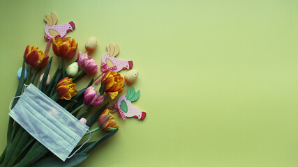 Easter eggs in pastel colors and fresh delicate tulips are gathered in a bouquet on a yellow background. Medical mask as a reminder of safety measures due to the covid-19 coronavirus pandemic.