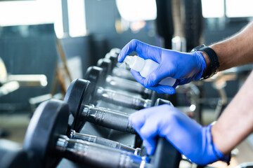 close up shot of hands with gloves sanitizing dumbbells at gym due to coronavirus - concept of covid-19 disinfecting, protection from virual infection at fitness