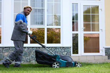 Smiling African man in overalls mows the green grass in a modern garden with a lawn mower. A black...