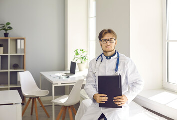 Portrait of confident young male doctor standing with clipboard in his hands in office of modern...