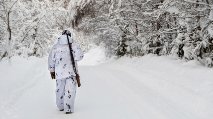 A male hunter in camouflage, armed with a rifle, stands with his back in a snowy winter forest.