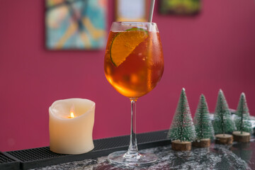 Aperol spritz summer cocktail in mister glass with lime, eco friendly straw and bubbles. Close up. Pink background. Toy Christmas trees, candle. 