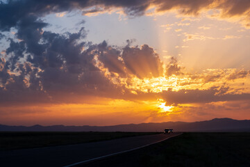 Beautiful sunset with cloudy sky and sun rays. Sunrise with road, mountains silhouettes and orange golden clouds.