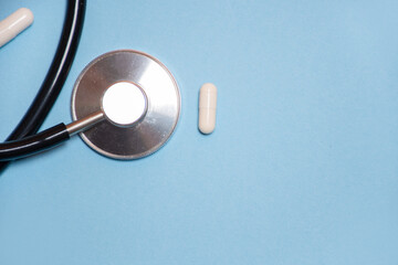 stethoscope or phonendoscope lies next to white tablets or capsules on a blue background: a place for text, high blood pressure, hypertension or hypotension and pulse diagnosis, crop image
