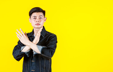 Portrait studio shot of Asian unsatisfied young male model in casual jeans jacket look at camera showing crossed hands symbol sign say no reject refuse deny on yellow background with copy space