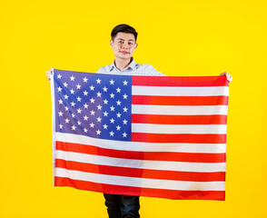 Portrait studio shot of millennial Asian young handsome male US citizen businessman model in casual outfit standing holding big United States of America USA national flag sheet on yellow background