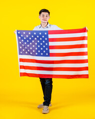 Portrait studio shot of millennial Asian young handsome male US citizen businessman model in casual outfit standing holding big United States of America USA national flag sheet on yellow background