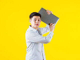 Portrait studio shot of millennial Asian young male businessman in casual outfit standing smiling holding laptop computer on yellow background