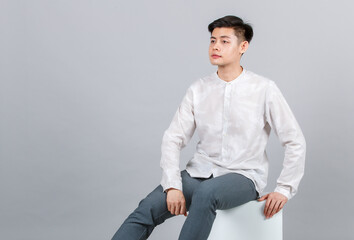 Studio shot of millennial Asian handsome young male model in casual outfit sitting on white square stool look at camera on gray background