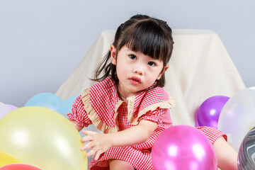 Portrait studio shot of little cute kindergarten preschooler kid girl daughter in red long dress sitting smiling alone on floor playing with colorful helium air party balloons on gray background