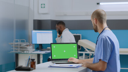 Obraz na płótnie Canvas Therapist man nurse sitting at desk typing sickness expertise on mock up green screen chroma key laptop computer with isolated display. Multi-ethnic team working at healthcare treatment