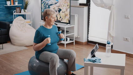 Elder adult following online training lesson on digital tablet. Senior woman using dumbbells to do physical activity and sitting on fitness toning ball while watching workout video on device.