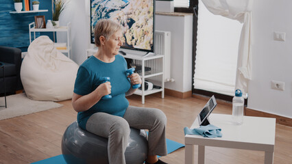 Elder adult following online training lesson on digital tablet. Senior woman using dumbbells to do...