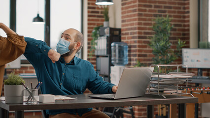 Business man greeting woman with elbow during covid 19 pandemic in startup office. Entrepreneur working with laptop while wearing face mask and talking to colleague. People at work