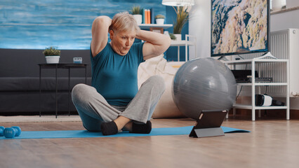 Elder person doing physical exercise and watching online lesson on tablet to workout. Retired woman...